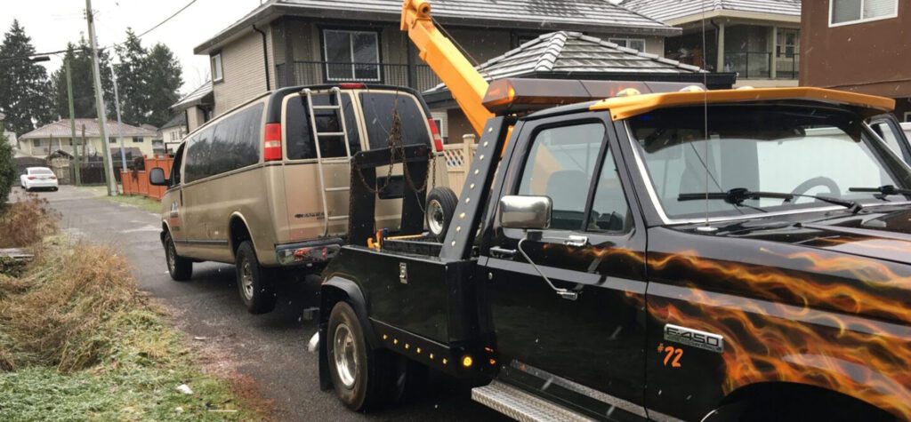 A tow truck with flame decals, offering Scrap Car Removal & Cash for Cars, is towing a silver van on a residential street with light snowfall.