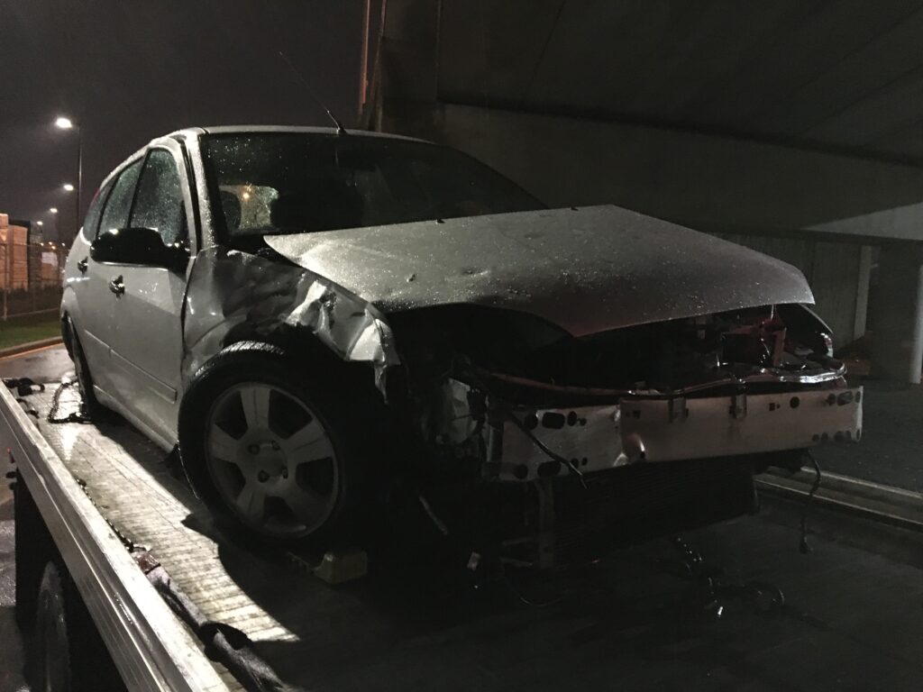 A damaged car with its front end crushed on a tow truck at night, awaiting scrap car removal.