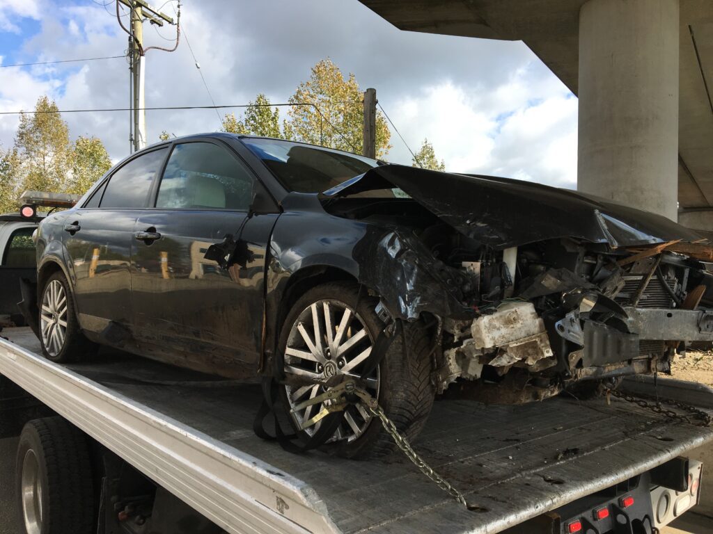 Damaged car with severe rear-end collision damage being towed on a flatbed truck for scrap car removal.