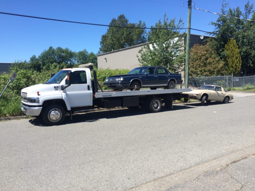 Flatbed tow truck carrying two vehicles for scrap car removal.